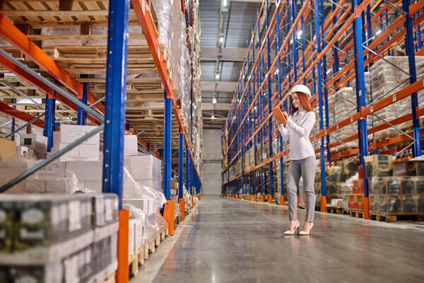 Our Services Include a young women taking stock check in warehouse with orange and blue racking Freight forwarding